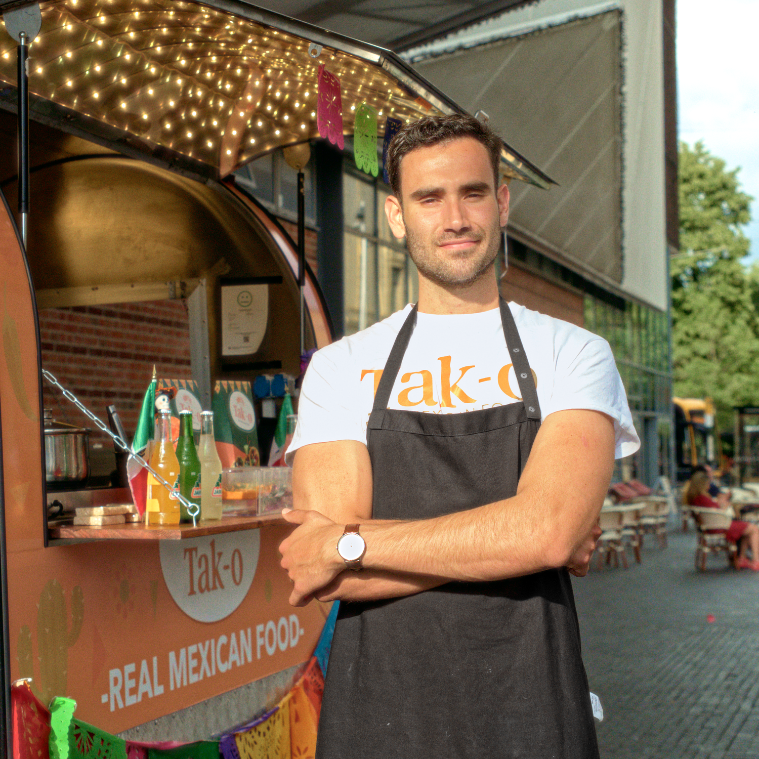 Man standing in front of a 'Tak-o' food truck, wearing a Nordgreen watch, white shirt and black apron, with arms crossed.